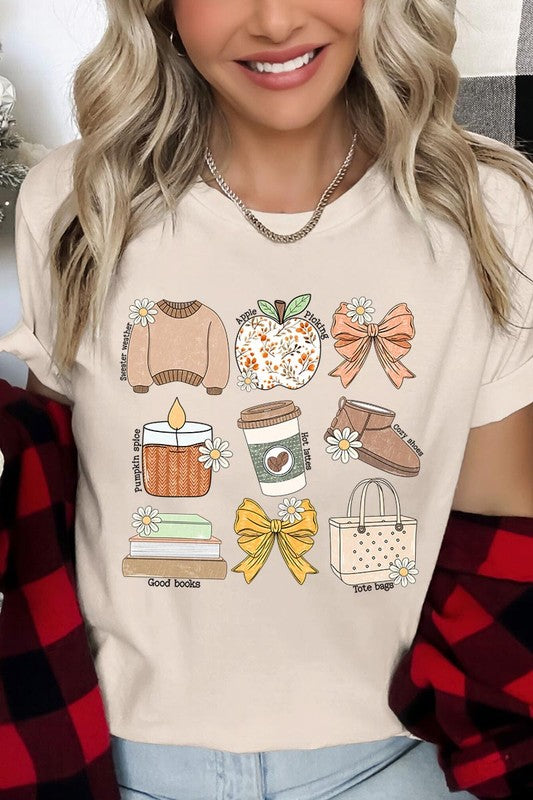 A person in a grey **Fall Girly Doodle Graphic Tee**, featuring charming illustrations of a sweater, apple, books, bows, coffee cup, candle, and tote bag, holds an orange mug while wearing a red cardigan and ripped jeans.