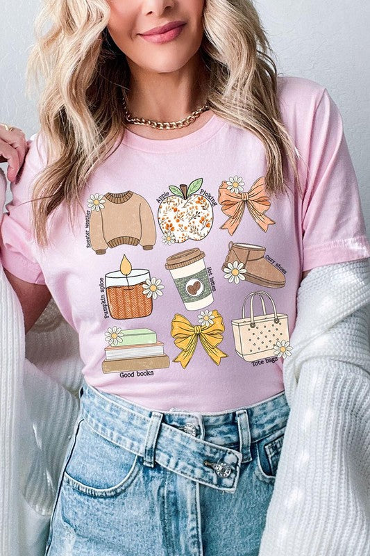 A person in a grey **Fall Girly Doodle Graphic Tee**, featuring charming illustrations of a sweater, apple, books, bows, coffee cup, candle, and tote bag, holds an orange mug while wearing a red cardigan and ripped jeans.