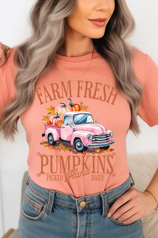 A person with blonde hair is wearing a black "Farm Fresh Pumpkins Truck Graphic Tee" that features a pink truck with pumpkins. They are holding a pumpkin and sitting indoors.