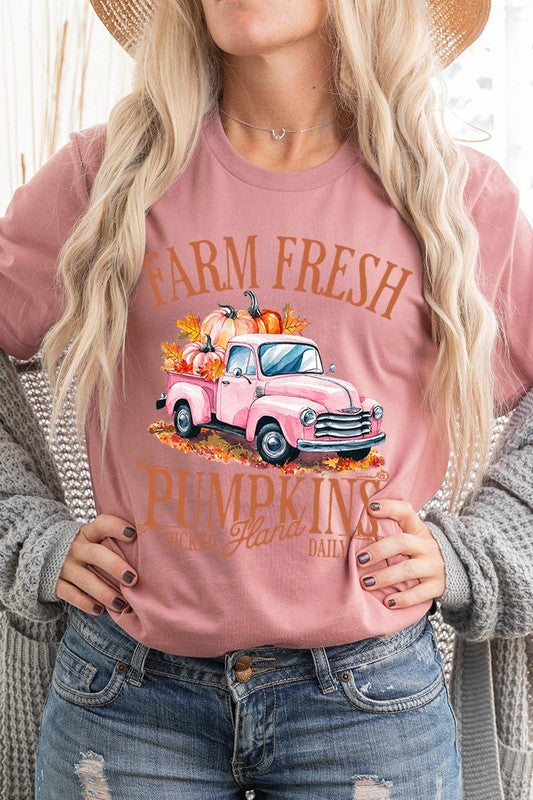 A person with blonde hair is wearing a black "Farm Fresh Pumpkins Truck Graphic Tee" that features a pink truck with pumpkins. They are holding a pumpkin and sitting indoors.