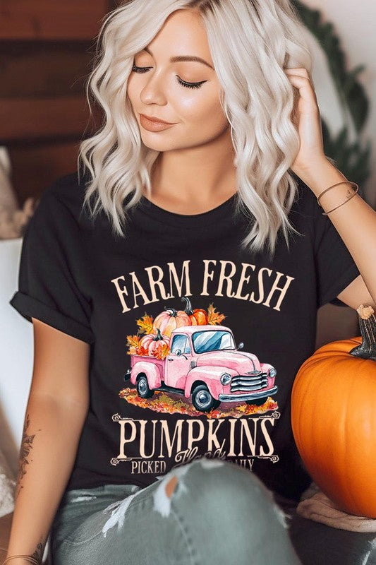 A person with blonde hair is wearing a black "Farm Fresh Pumpkins Truck Graphic Tee" that features a pink truck with pumpkins. They are holding a pumpkin and sitting indoors.