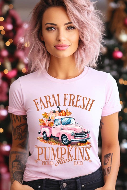 A person with blonde hair is wearing a black "Farm Fresh Pumpkins Truck Graphic Tee" that features a pink truck with pumpkins. They are holding a pumpkin and sitting indoors.