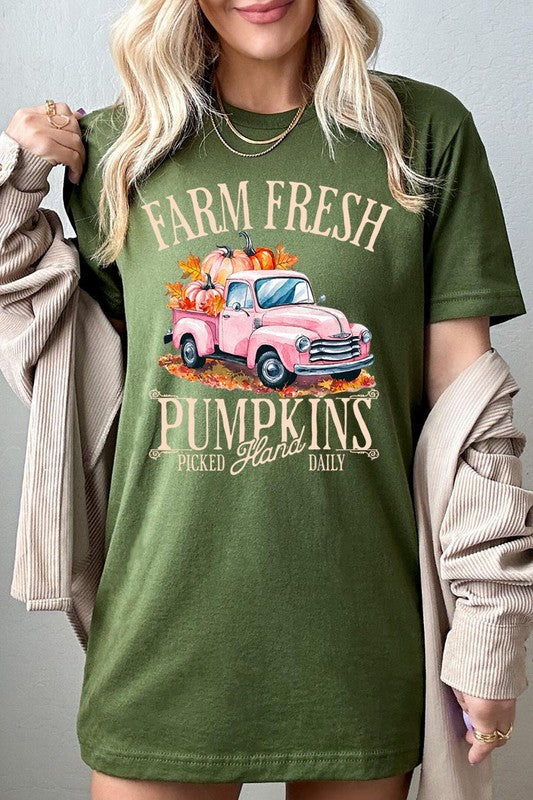 A person with blonde hair is wearing a black "Farm Fresh Pumpkins Truck Graphic Tee" that features a pink truck with pumpkins. They are holding a pumpkin and sitting indoors.