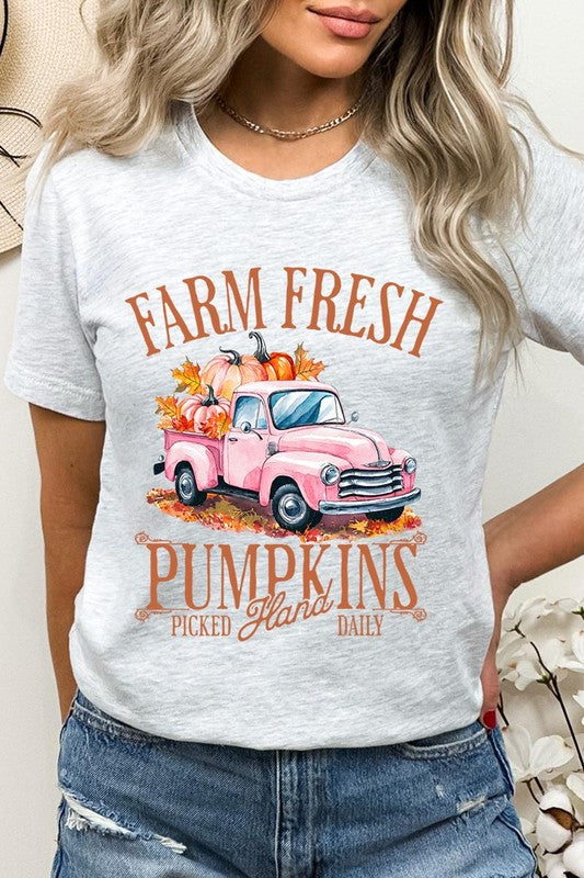 A person with blonde hair is wearing a black "Farm Fresh Pumpkins Truck Graphic Tee" that features a pink truck with pumpkins. They are holding a pumpkin and sitting indoors.