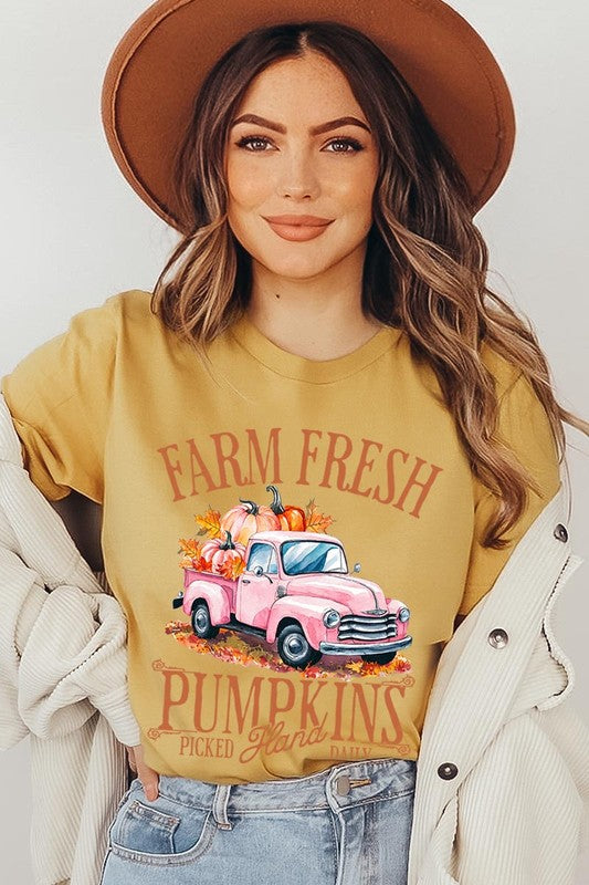 A person with blonde hair is wearing a black "Farm Fresh Pumpkins Truck Graphic Tee" that features a pink truck with pumpkins. They are holding a pumpkin and sitting indoors.