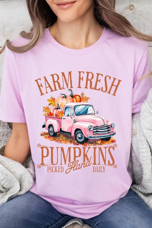 A person with blonde hair is wearing a black "Farm Fresh Pumpkins Truck Graphic Tee" that features a pink truck with pumpkins. They are holding a pumpkin and sitting indoors.