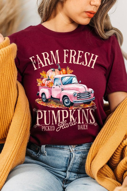 A person with blonde hair is wearing a black "Farm Fresh Pumpkins Truck Graphic Tee" that features a pink truck with pumpkins. They are holding a pumpkin and sitting indoors.