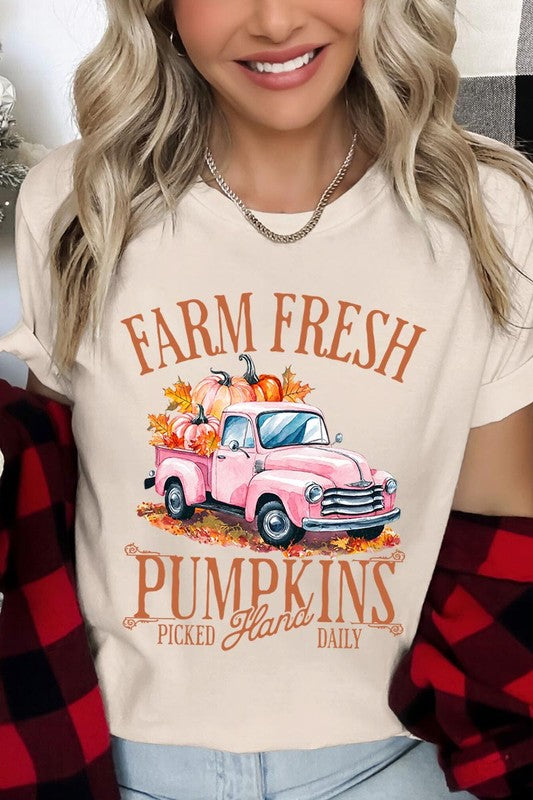 A person with blonde hair is wearing a black "Farm Fresh Pumpkins Truck Graphic Tee" that features a pink truck with pumpkins. They are holding a pumpkin and sitting indoors.