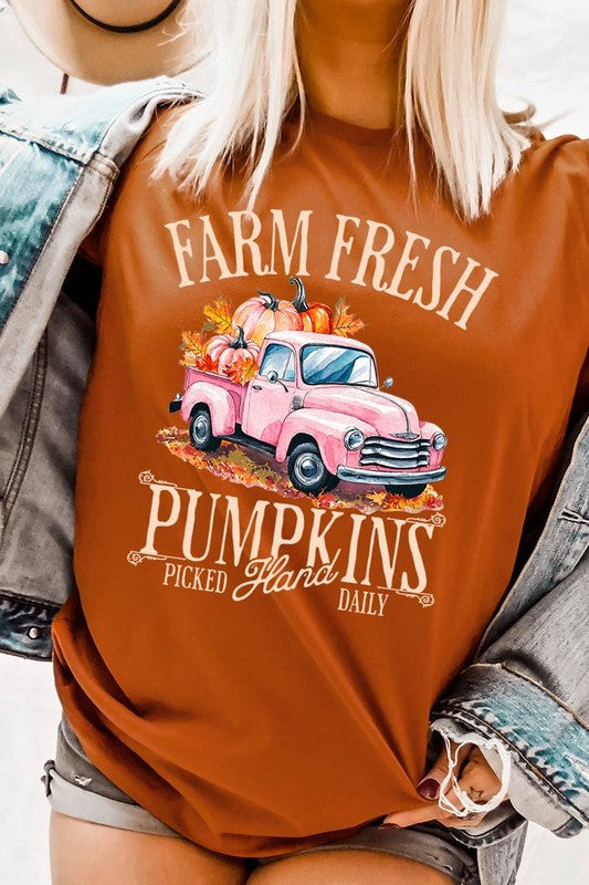 A person with blonde hair is wearing a black "Farm Fresh Pumpkins Truck Graphic Tee" that features a pink truck with pumpkins. They are holding a pumpkin and sitting indoors.