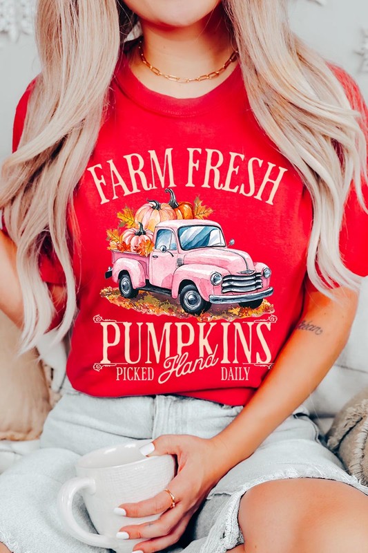 A person with blonde hair is wearing a black "Farm Fresh Pumpkins Truck Graphic Tee" that features a pink truck with pumpkins. They are holding a pumpkin and sitting indoors.