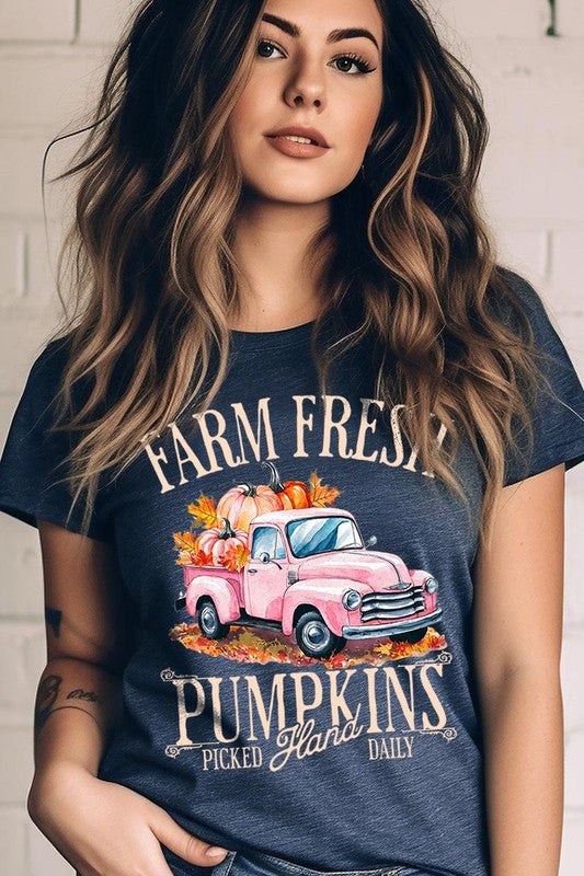 A person with blonde hair is wearing a black "Farm Fresh Pumpkins Truck Graphic Tee" that features a pink truck with pumpkins. They are holding a pumpkin and sitting indoors.