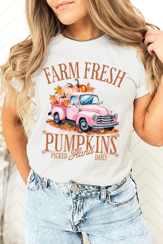 A person with blonde hair is wearing a black "Farm Fresh Pumpkins Truck Graphic Tee" that features a pink truck with pumpkins. They are holding a pumpkin and sitting indoors.