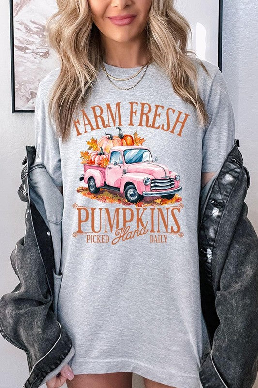 A person with blonde hair is wearing a black "Farm Fresh Pumpkins Truck Graphic Tee" that features a pink truck with pumpkins. They are holding a pumpkin and sitting indoors.