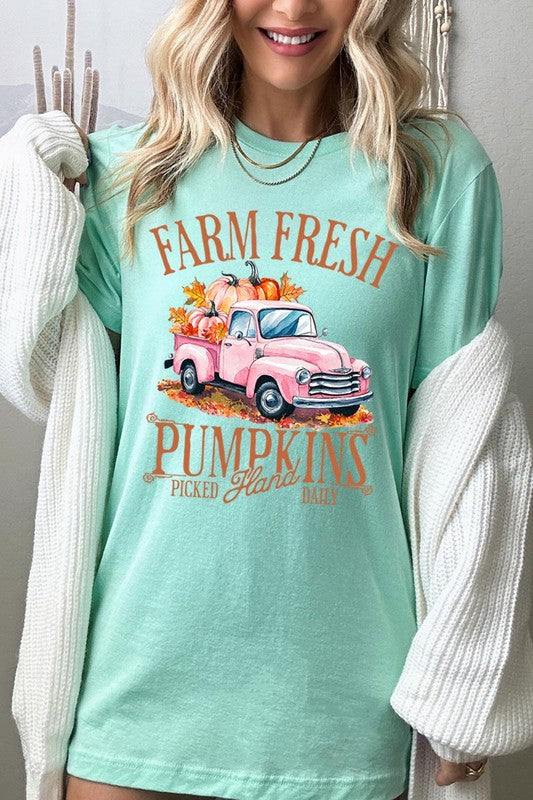 A person with blonde hair is wearing a black "Farm Fresh Pumpkins Truck Graphic Tee" that features a pink truck with pumpkins. They are holding a pumpkin and sitting indoors.