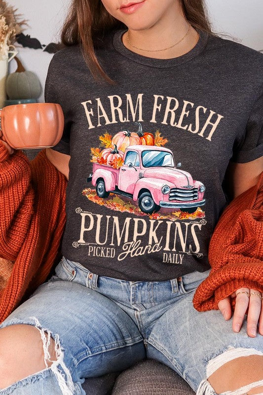 A person with blonde hair is wearing a black "Farm Fresh Pumpkins Truck Graphic Tee" that features a pink truck with pumpkins. They are holding a pumpkin and sitting indoors.