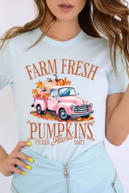 A person with blonde hair is wearing a black "Farm Fresh Pumpkins Truck Graphic Tee" that features a pink truck with pumpkins. They are holding a pumpkin and sitting indoors.