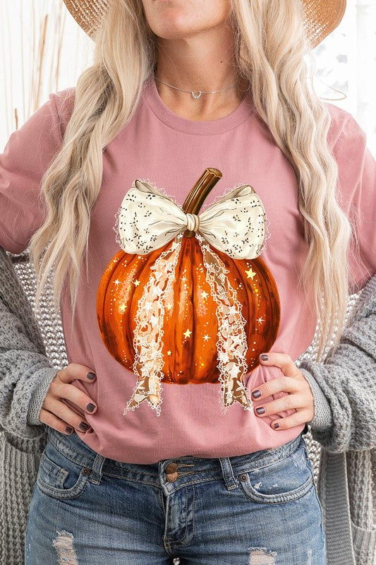 A person is wearing a Coquette bow Halloween Pumpkin Graphic Tee, which is a beige unisex crew neck shirt featuring a large illustrated Halloween pumpkin adorned with a white lace bow and sparkles on the front. The pumpkin has a vertical wooden stem. The person also has on a denim jacket.