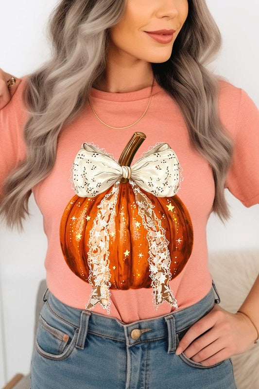 A person is wearing a Coquette bow Halloween Pumpkin Graphic Tee, which is a beige unisex crew neck shirt featuring a large illustrated Halloween pumpkin adorned with a white lace bow and sparkles on the front. The pumpkin has a vertical wooden stem. The person also has on a denim jacket.