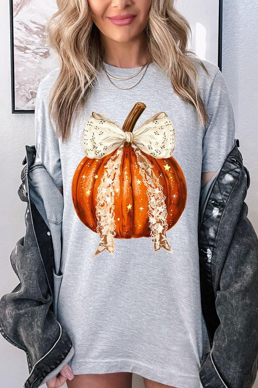 A person is wearing a Coquette bow Halloween Pumpkin Graphic Tee, which is a beige unisex crew neck shirt featuring a large illustrated Halloween pumpkin adorned with a white lace bow and sparkles on the front. The pumpkin has a vertical wooden stem. The person also has on a denim jacket.