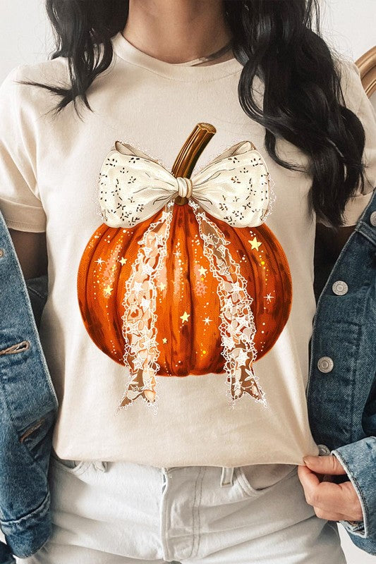 A person is wearing a Coquette bow Halloween Pumpkin Graphic Tee, which is a beige unisex crew neck shirt featuring a large illustrated Halloween pumpkin adorned with a white lace bow and sparkles on the front. The pumpkin has a vertical wooden stem. The person also has on a denim jacket.