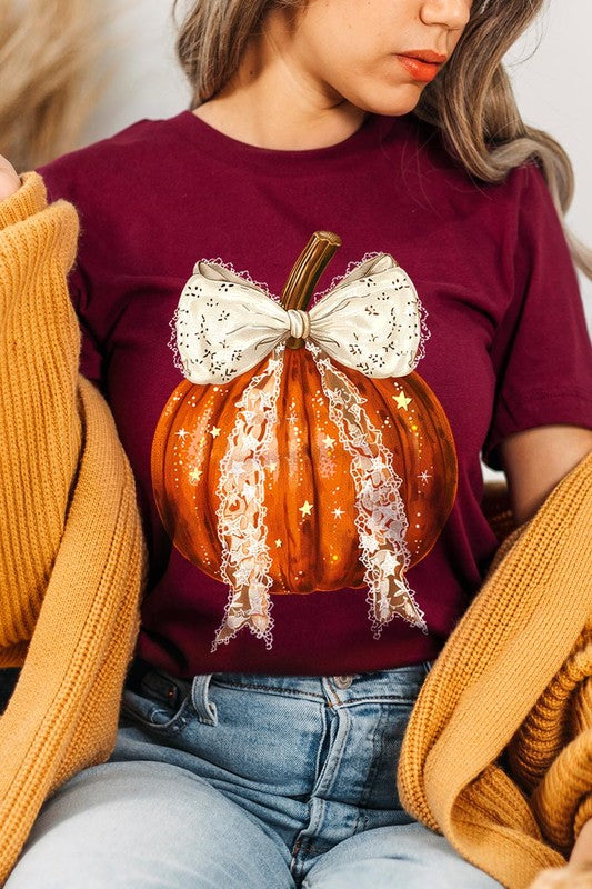 A person is wearing a Coquette bow Halloween Pumpkin Graphic Tee, which is a beige unisex crew neck shirt featuring a large illustrated Halloween pumpkin adorned with a white lace bow and sparkles on the front. The pumpkin has a vertical wooden stem. The person also has on a denim jacket.