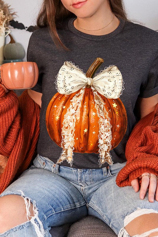 A person is wearing a Coquette bow Halloween Pumpkin Graphic Tee, which is a beige unisex crew neck shirt featuring a large illustrated Halloween pumpkin adorned with a white lace bow and sparkles on the front. The pumpkin has a vertical wooden stem. The person also has on a denim jacket.