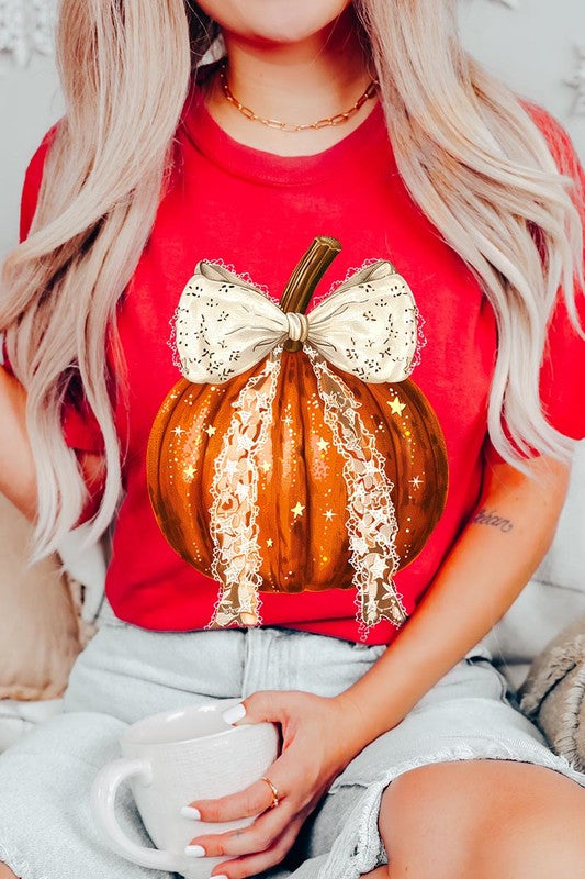 A person is wearing a Coquette bow Halloween Pumpkin Graphic Tee, which is a beige unisex crew neck shirt featuring a large illustrated Halloween pumpkin adorned with a white lace bow and sparkles on the front. The pumpkin has a vertical wooden stem. The person also has on a denim jacket.