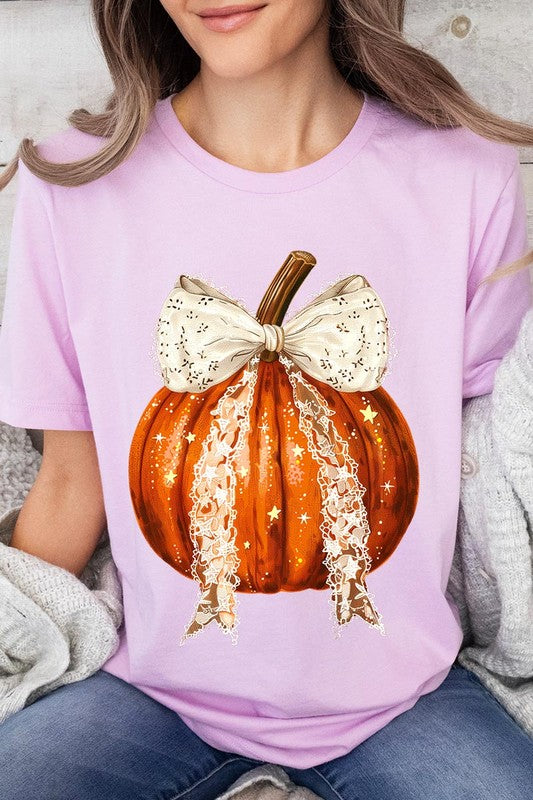 A person is wearing a Coquette bow Halloween Pumpkin Graphic Tee, which is a beige unisex crew neck shirt featuring a large illustrated Halloween pumpkin adorned with a white lace bow and sparkles on the front. The pumpkin has a vertical wooden stem. The person also has on a denim jacket.