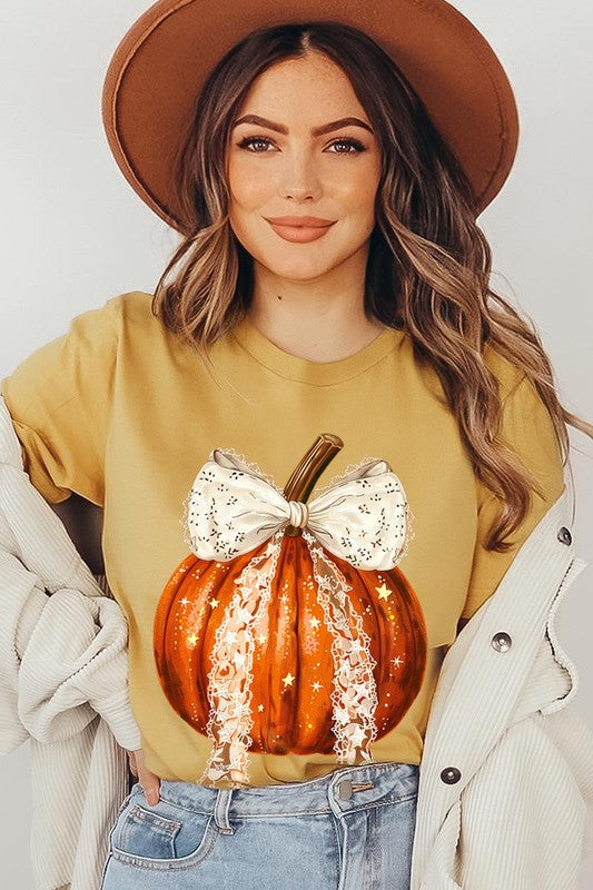 A person is wearing a Coquette bow Halloween Pumpkin Graphic Tee, which is a beige unisex crew neck shirt featuring a large illustrated Halloween pumpkin adorned with a white lace bow and sparkles on the front. The pumpkin has a vertical wooden stem. The person also has on a denim jacket.