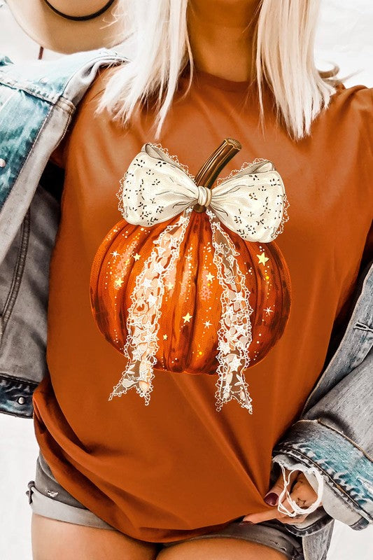 A person is wearing a Coquette bow Halloween Pumpkin Graphic Tee, which is a beige unisex crew neck shirt featuring a large illustrated Halloween pumpkin adorned with a white lace bow and sparkles on the front. The pumpkin has a vertical wooden stem. The person also has on a denim jacket.