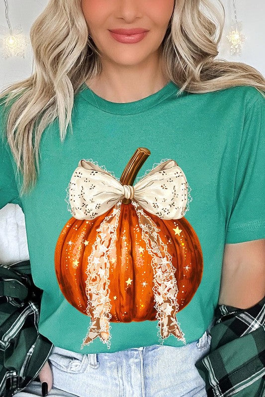 A person is wearing a Coquette bow Halloween Pumpkin Graphic Tee, which is a beige unisex crew neck shirt featuring a large illustrated Halloween pumpkin adorned with a white lace bow and sparkles on the front. The pumpkin has a vertical wooden stem. The person also has on a denim jacket.