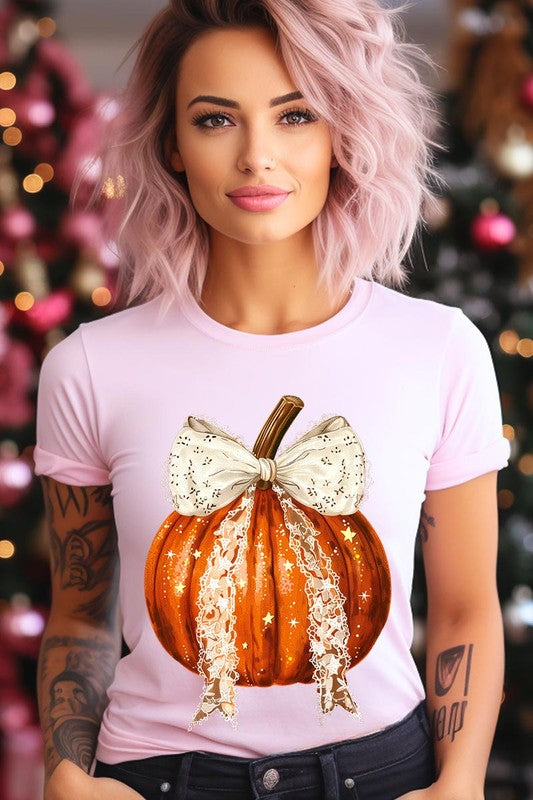 A person is wearing a Coquette bow Halloween Pumpkin Graphic Tee, which is a beige unisex crew neck shirt featuring a large illustrated Halloween pumpkin adorned with a white lace bow and sparkles on the front. The pumpkin has a vertical wooden stem. The person also has on a denim jacket.