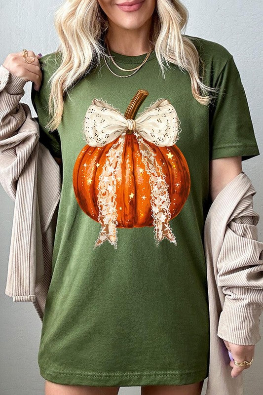 A person is wearing a Coquette bow Halloween Pumpkin Graphic Tee, which is a beige unisex crew neck shirt featuring a large illustrated Halloween pumpkin adorned with a white lace bow and sparkles on the front. The pumpkin has a vertical wooden stem. The person also has on a denim jacket.