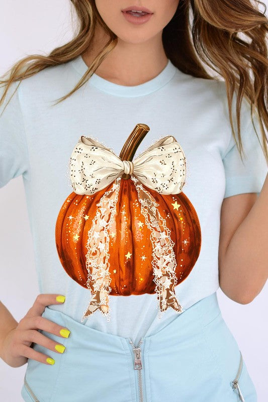 A person is wearing a Coquette bow Halloween Pumpkin Graphic Tee, which is a beige unisex crew neck shirt featuring a large illustrated Halloween pumpkin adorned with a white lace bow and sparkles on the front. The pumpkin has a vertical wooden stem. The person also has on a denim jacket.