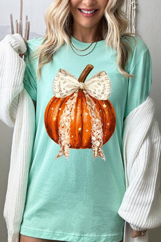 A person is wearing a Coquette bow Halloween Pumpkin Graphic Tee, which is a beige unisex crew neck shirt featuring a large illustrated Halloween pumpkin adorned with a white lace bow and sparkles on the front. The pumpkin has a vertical wooden stem. The person also has on a denim jacket.