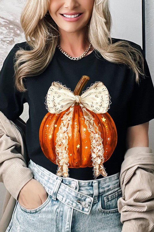 A person is wearing a Coquette bow Halloween Pumpkin Graphic Tee, which is a beige unisex crew neck shirt featuring a large illustrated Halloween pumpkin adorned with a white lace bow and sparkles on the front. The pumpkin has a vertical wooden stem. The person also has on a denim jacket.