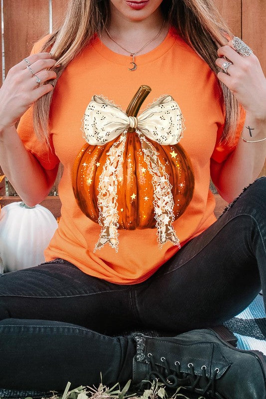 A person is wearing a Coquette bow Halloween Pumpkin Graphic Tee, which is a beige unisex crew neck shirt featuring a large illustrated Halloween pumpkin adorned with a white lace bow and sparkles on the front. The pumpkin has a vertical wooden stem. The person also has on a denim jacket.