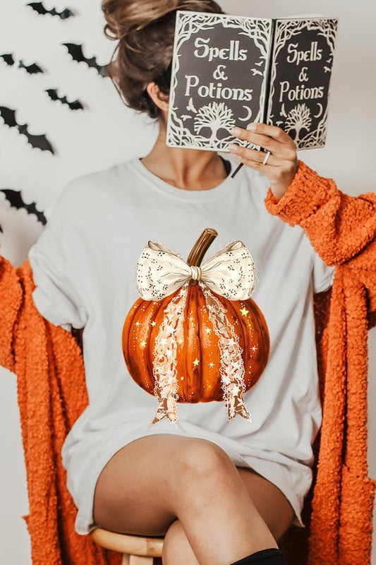 A person is wearing a Coquette bow Halloween Pumpkin Graphic Tee, which is a beige unisex crew neck shirt featuring a large illustrated Halloween pumpkin adorned with a white lace bow and sparkles on the front. The pumpkin has a vertical wooden stem. The person also has on a denim jacket.