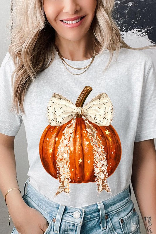 A person is wearing a Coquette bow Halloween Pumpkin Graphic Tee, which is a beige unisex crew neck shirt featuring a large illustrated Halloween pumpkin adorned with a white lace bow and sparkles on the front. The pumpkin has a vertical wooden stem. The person also has on a denim jacket.