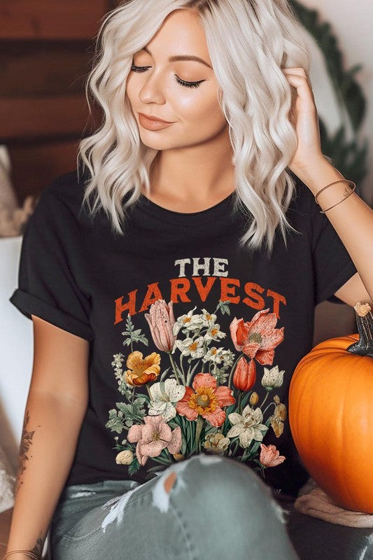 A woman is pictured wearing The Harvest Christian Graphic Tee, which features "The Harvest" in bold letters, an illustration of various flowers, and the verse "Is plentiful, but the laborers are few. Matthew 9:37." She has her denim jacket open to display the tee. This versatile shirt is ideal for Family Group Uniforms or as a Unisex Crew Neck option.