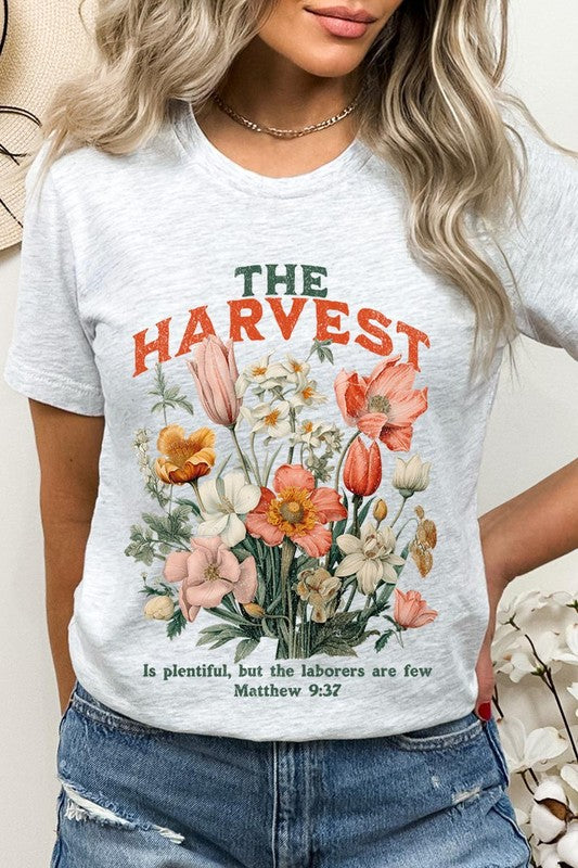 A woman is pictured wearing The Harvest Christian Graphic Tee, which features "The Harvest" in bold letters, an illustration of various flowers, and the verse "Is plentiful, but the laborers are few. Matthew 9:37." She has her denim jacket open to display the tee. This versatile shirt is ideal for Family Group Uniforms or as a Unisex Crew Neck option.