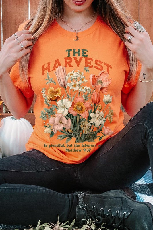 A woman is pictured wearing The Harvest Christian Graphic Tee, which features "The Harvest" in bold letters, an illustration of various flowers, and the verse "Is plentiful, but the laborers are few. Matthew 9:37." She has her denim jacket open to display the tee. This versatile shirt is ideal for Family Group Uniforms or as a Unisex Crew Neck option.