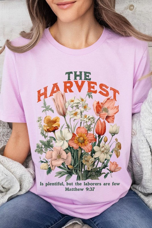 A woman is pictured wearing The Harvest Christian Graphic Tee, which features "The Harvest" in bold letters, an illustration of various flowers, and the verse "Is plentiful, but the laborers are few. Matthew 9:37." She has her denim jacket open to display the tee. This versatile shirt is ideal for Family Group Uniforms or as a Unisex Crew Neck option.