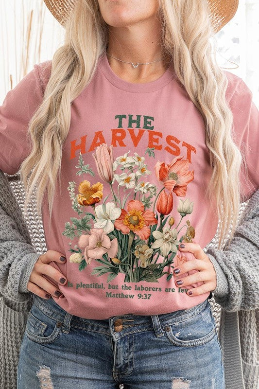 A woman is pictured wearing The Harvest Christian Graphic Tee, which features "The Harvest" in bold letters, an illustration of various flowers, and the verse "Is plentiful, but the laborers are few. Matthew 9:37." She has her denim jacket open to display the tee. This versatile shirt is ideal for Family Group Uniforms or as a Unisex Crew Neck option.