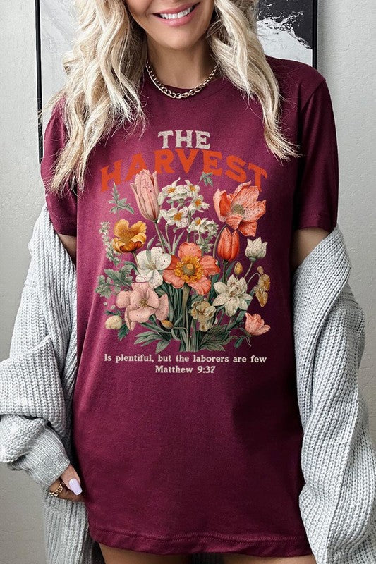 A woman is pictured wearing The Harvest Christian Graphic Tee, which features "The Harvest" in bold letters, an illustration of various flowers, and the verse "Is plentiful, but the laborers are few. Matthew 9:37." She has her denim jacket open to display the tee. This versatile shirt is ideal for Family Group Uniforms or as a Unisex Crew Neck option.