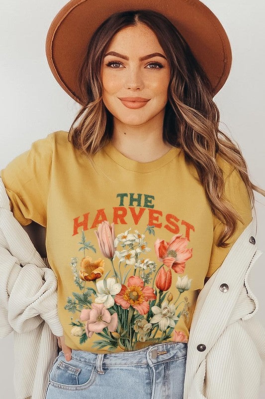 A woman is pictured wearing The Harvest Christian Graphic Tee, which features "The Harvest" in bold letters, an illustration of various flowers, and the verse "Is plentiful, but the laborers are few. Matthew 9:37." She has her denim jacket open to display the tee. This versatile shirt is ideal for Family Group Uniforms or as a Unisex Crew Neck option.