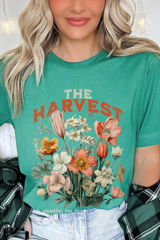 A woman is pictured wearing The Harvest Christian Graphic Tee, which features "The Harvest" in bold letters, an illustration of various flowers, and the verse "Is plentiful, but the laborers are few. Matthew 9:37." She has her denim jacket open to display the tee. This versatile shirt is ideal for Family Group Uniforms or as a Unisex Crew Neck option.