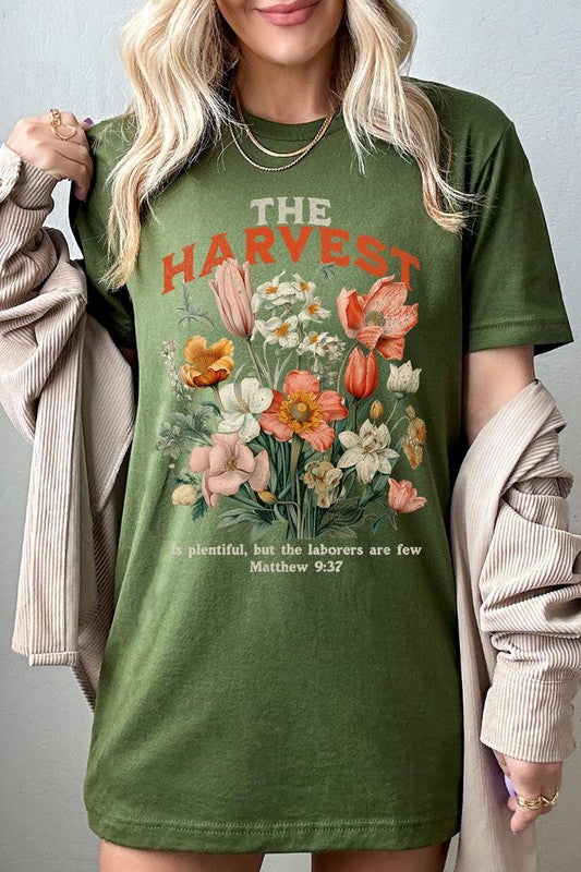 A woman is pictured wearing The Harvest Christian Graphic Tee, which features "The Harvest" in bold letters, an illustration of various flowers, and the verse "Is plentiful, but the laborers are few. Matthew 9:37." She has her denim jacket open to display the tee. This versatile shirt is ideal for Family Group Uniforms or as a Unisex Crew Neck option.