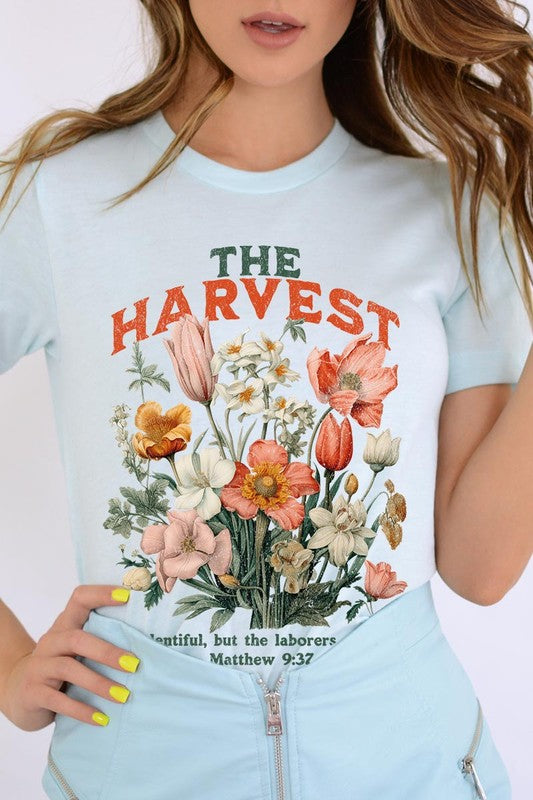 A woman is pictured wearing The Harvest Christian Graphic Tee, which features "The Harvest" in bold letters, an illustration of various flowers, and the verse "Is plentiful, but the laborers are few. Matthew 9:37." She has her denim jacket open to display the tee. This versatile shirt is ideal for Family Group Uniforms or as a Unisex Crew Neck option.