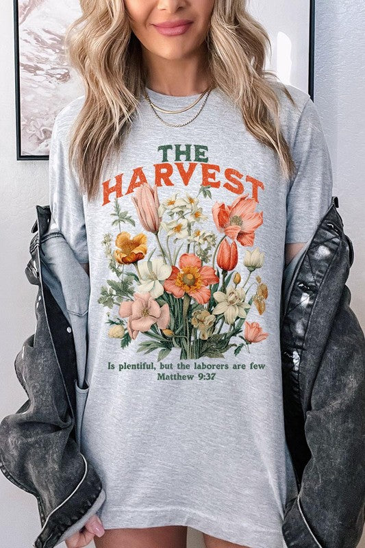 A woman is pictured wearing The Harvest Christian Graphic Tee, which features "The Harvest" in bold letters, an illustration of various flowers, and the verse "Is plentiful, but the laborers are few. Matthew 9:37." She has her denim jacket open to display the tee. This versatile shirt is ideal for Family Group Uniforms or as a Unisex Crew Neck option.